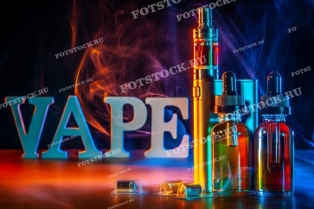 На фотографии изображены буквы, составляющие слово "VAPE", и различные аксессуары для вейпинга, такие как электронные устройства и флаконы с жидкостью, на фоне дыма. Все элементы расположены на деревянной поверхности.
